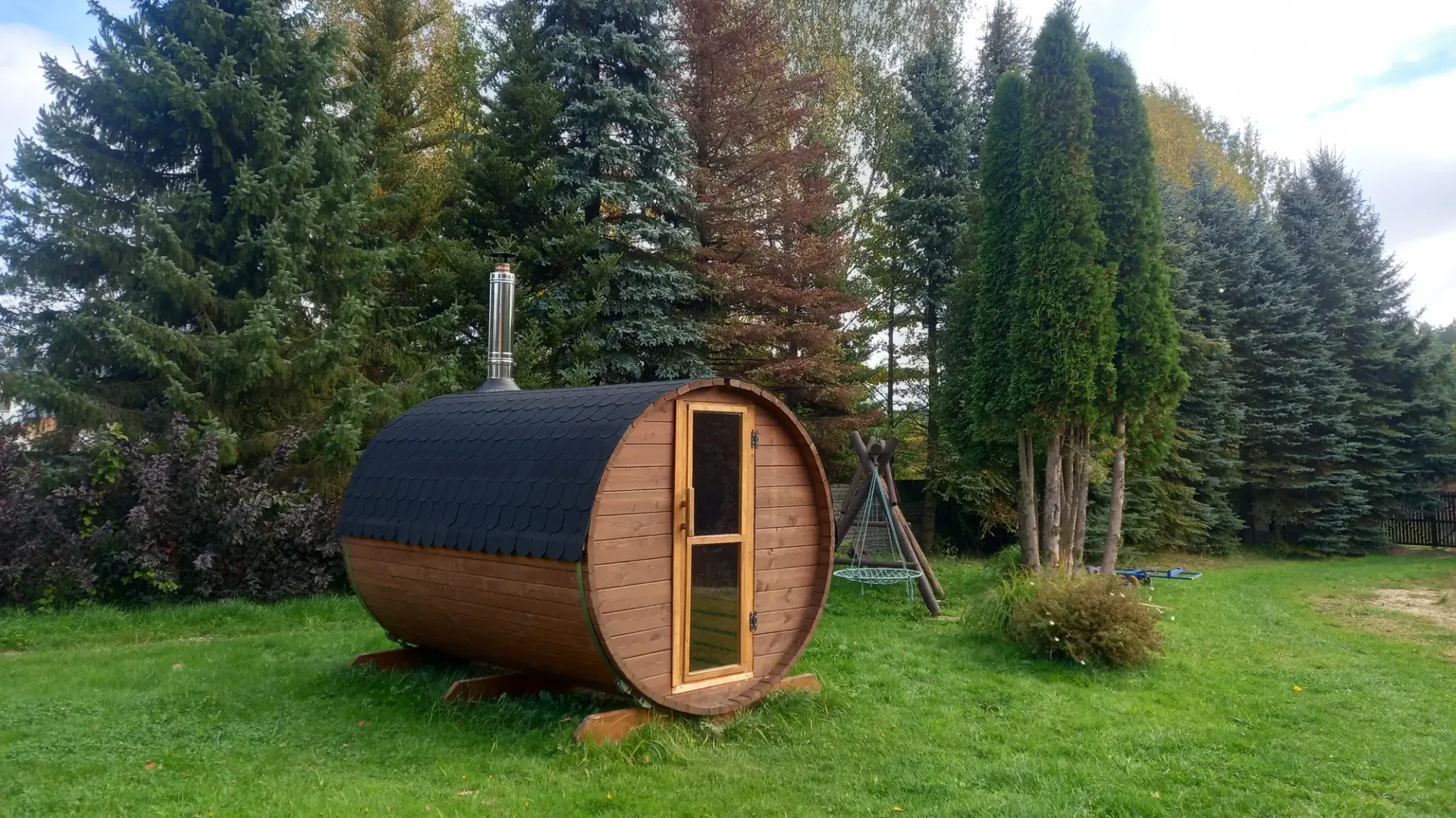 Sauna ogrodowa w kształcie beczki na trawniku wśród świerków