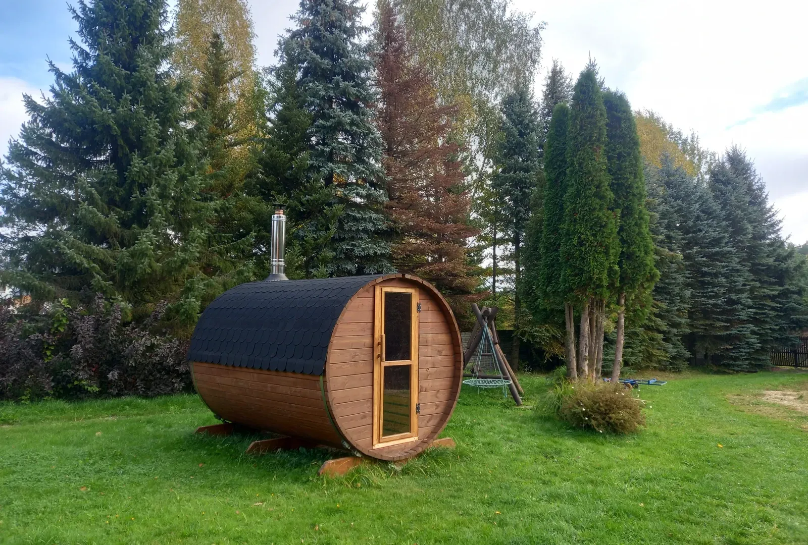 Drewniana sauna ogrodowa w kształcie beczki na trawniku wśród świerków i tui