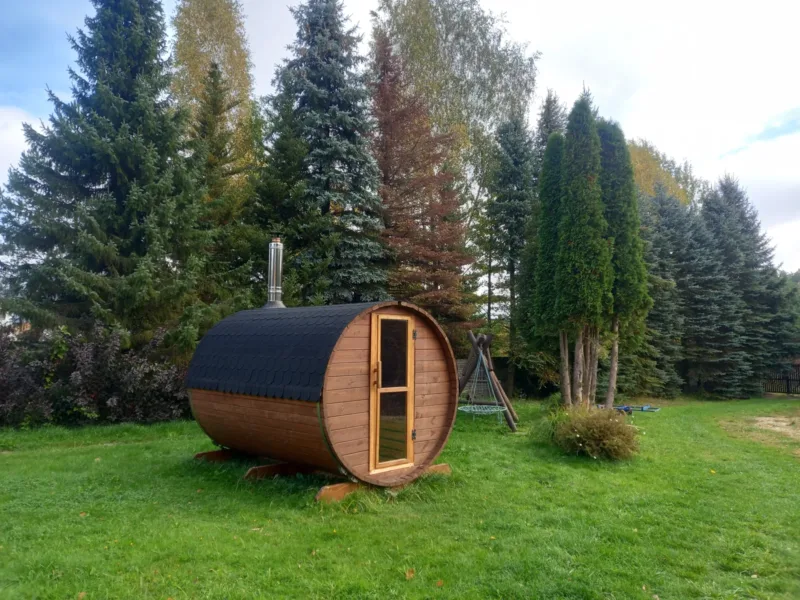 Drewniana sauna ogrodowa w kształcie beczki na trawniku wśród świerków i tui